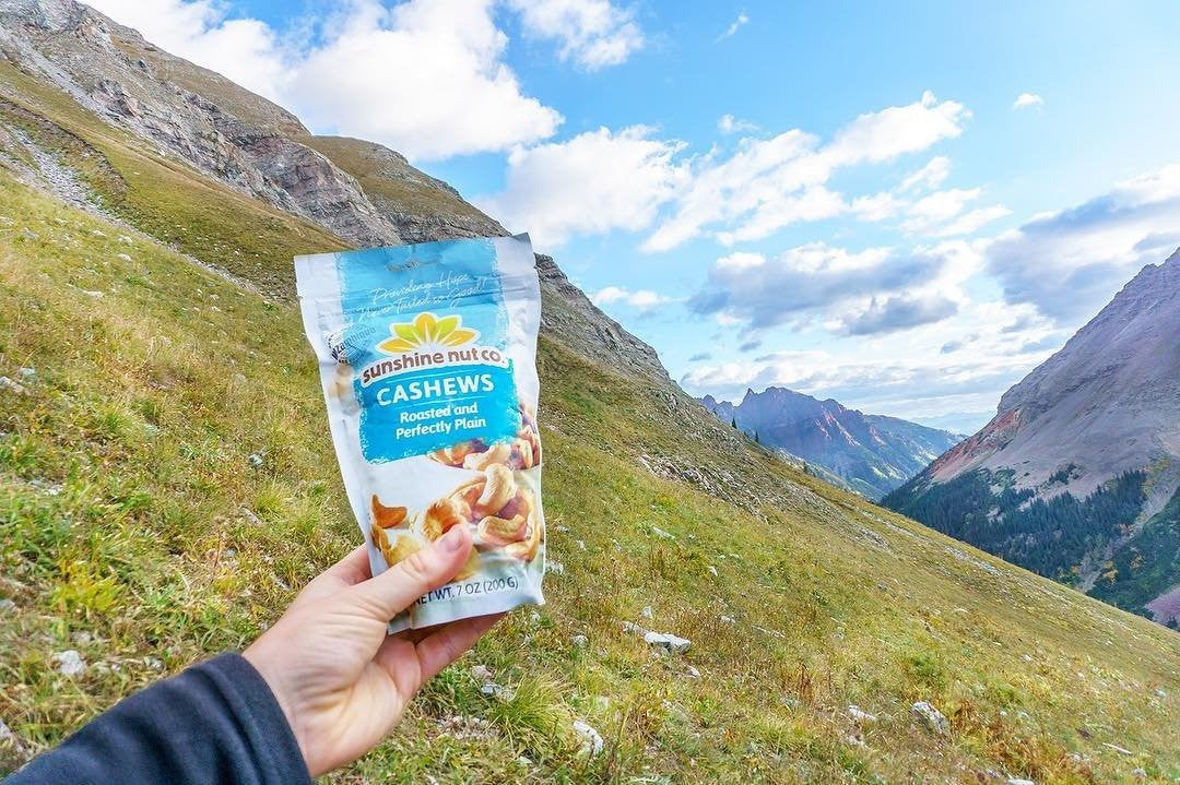 Handing Holding 7 Ounce Bag Of Roasted And Perfectly Plain Sunshine Nut Co. Cashews Outside On Mountain Hill